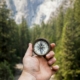 A hand holding an open compass in front of a pine forest and mountain cliffs, representing values as a direction in acceptance and commitment therapy