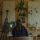 A person seated at a warmly lit desk in a plant-filled studio, working quietly with notes and artwork on the walls.