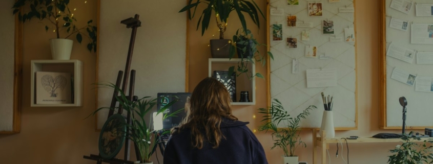 A person seated at a warmly lit desk in a plant-filled studio, working quietly with notes and artwork on the walls.