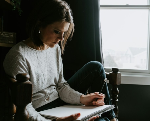 Woman sitting in a chair by a window writing in a notebook, keeping a daily mood log