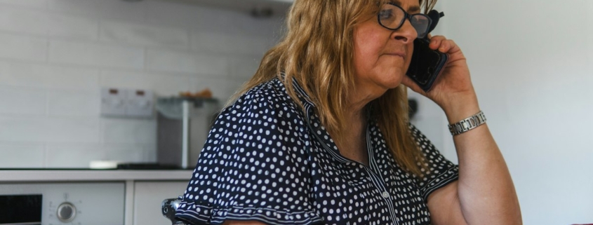 A woman sitting at her kitchen table making a phone call with a concerned expression