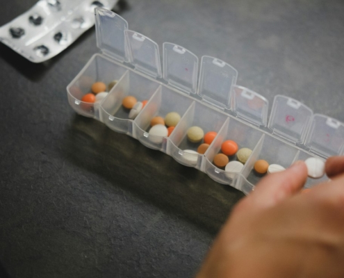 Hand taking a daily dose from a weekly pill organizer on a dark table, representing ongoing medical care alongside residential mental health treatment
