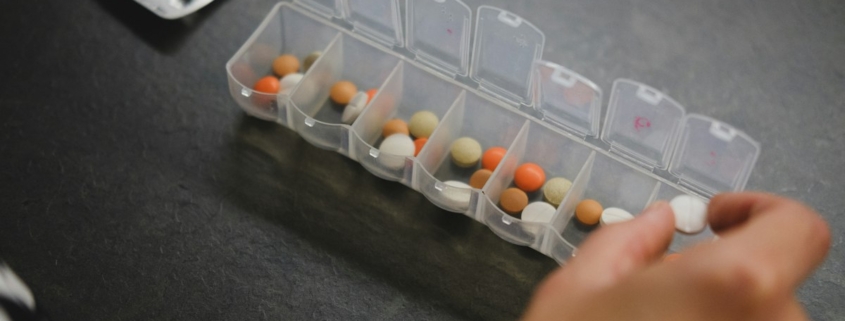 Hand taking a daily dose from a weekly pill organizer on a dark table, representing ongoing medical care alongside residential mental health treatment