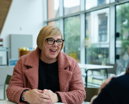 Two women talking at a table in a bright, window-lined room, one smiling as she listens to questions about a research study