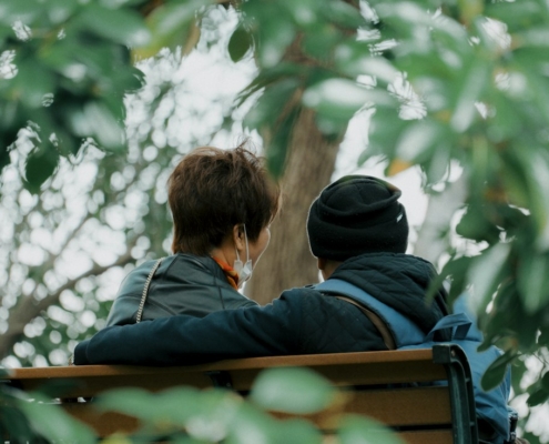 Two people sitting close together on a park bench under green leaves, one with an arm around the other