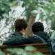 Two people sitting close together on a park bench under green leaves, one with an arm around the other