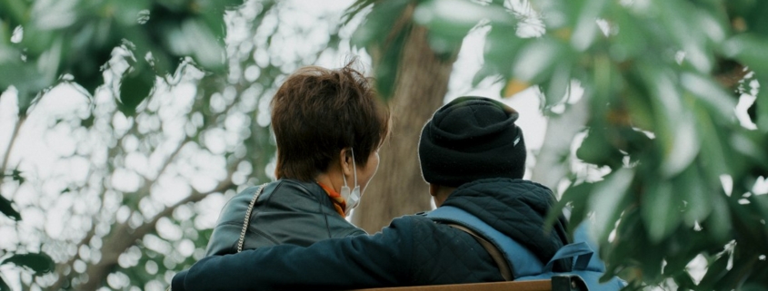 Two people sitting close together on a park bench under green leaves, one with an arm around the other