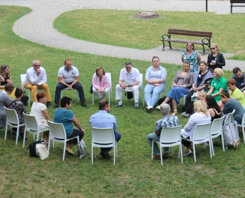 A group of adults seated in a circle of white chairs on a lawn, talking together outdoors