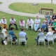 A group of adults seated in a circle of white chairs on a lawn, talking together outdoors