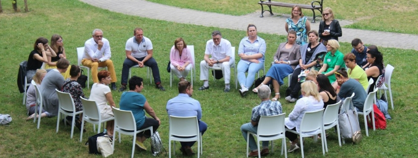 A group of adults seated in a circle of white chairs on a lawn, talking together outdoors