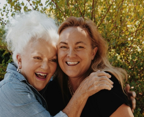 Adult daughter and her mother hugging and smiling outdoors in front of green shrubs