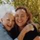 Adult daughter and her mother hugging and smiling outdoors in front of green shrubs