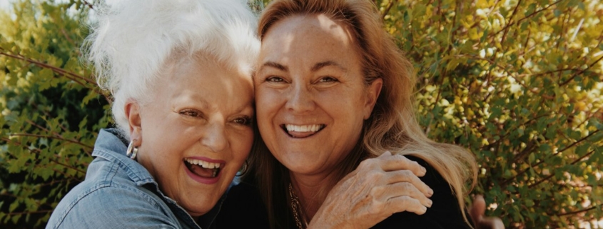 Adult daughter and her mother hugging and smiling outdoors in front of green shrubs