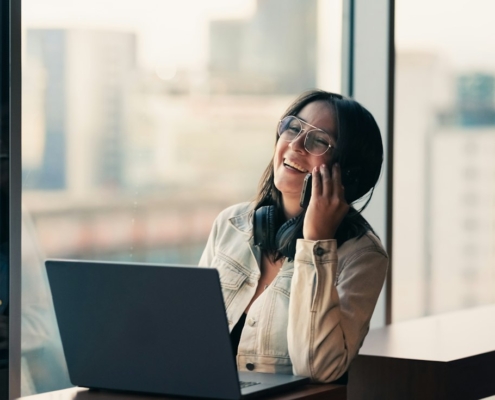 A woman smiling as she takes a phone call at a desk by a bright window