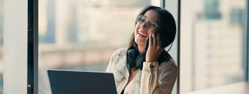 A woman smiling as she takes a phone call at a desk by a bright window