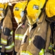 Rows of firefighter turnout coats and helmets hanging on a station rack in warm morning light