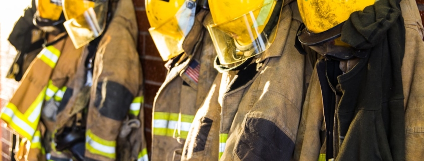 Rows of firefighter turnout coats and helmets hanging on a station rack in warm morning light