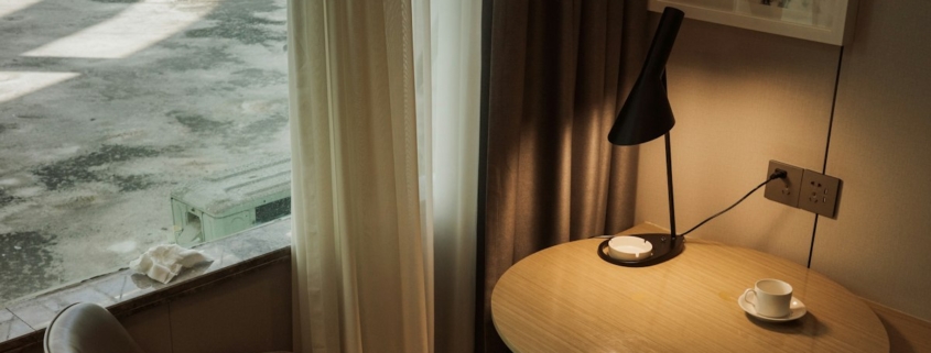 A quiet sunlit corner with an armchair, a small round table, a lamp and a cup beside a curtained window