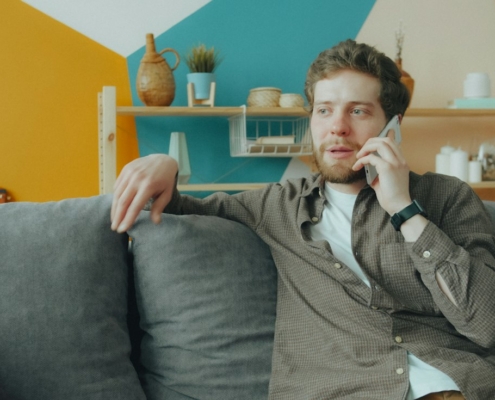 A man sitting on a gray sofa at home talking on his phone, calling about insurance coverage for residential mental health treatment