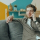 A man sitting on a gray sofa at home talking on his phone, calling about insurance coverage for residential mental health treatment