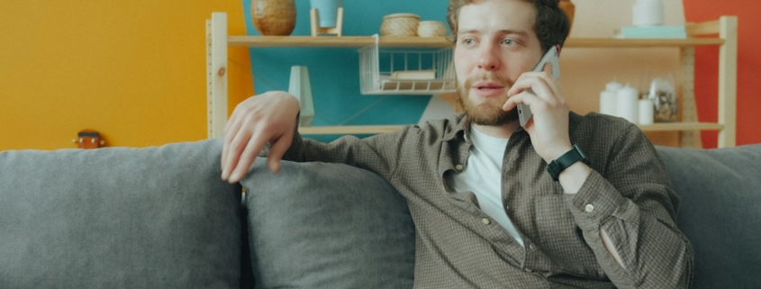 A man sitting on a gray sofa at home talking on his phone, calling about insurance coverage for residential mental health treatment