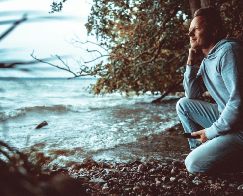 Man in a light hoodie crouching on a rocky shoreline, looking out calmly over the water