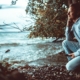 Man in a light hoodie crouching on a rocky shoreline, looking out calmly over the water
