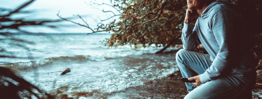 Man in a light hoodie crouching on a rocky shoreline, looking out calmly over the water