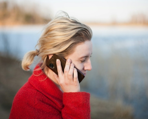 A woman in a red coat talking on her phone beside a calm lake in soft evening light