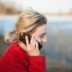 A woman in a red coat talking on her phone beside a calm lake in soft evening light