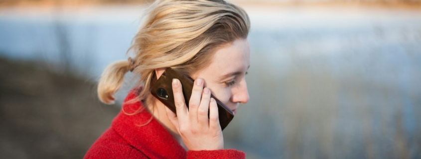 A woman in a red coat talking on her phone beside a calm lake in soft evening light