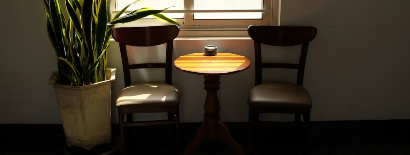 Two wooden chairs and a small round table beside a sunlit window, a calm setting for a conversation at home
