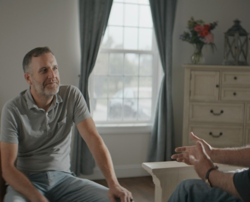 Two adults sitting and talking in a quiet, sunlit room, one listening while the other speaks