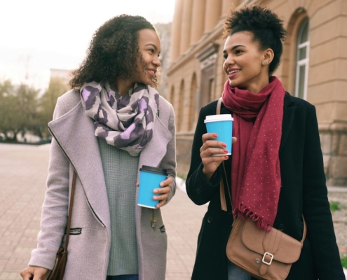 Two women walking and talking together outdoors, holding coffee cups, in a calm, supportive conversation
