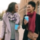 Two women walking and talking together outdoors, holding coffee cups, in a calm, supportive conversation