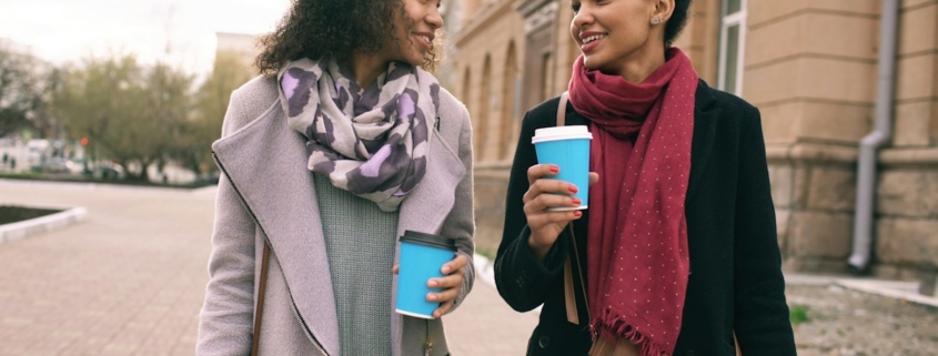 Two women walking and talking together outdoors, holding coffee cups, in a calm, supportive conversation