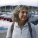 Woman in midlife standing outdoors near a harbor on a calm overcast morning, looking thoughtful