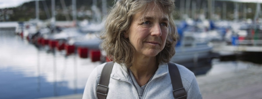 Woman in midlife standing outdoors near a harbor on a calm overcast morning, looking thoughtful