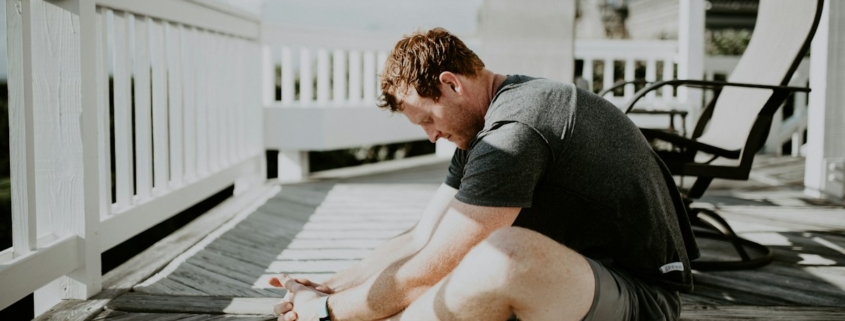 A man doing a seated forward stretch on a sunlit wooden porch, illustrating gentle physical activity during residential mental health treatment