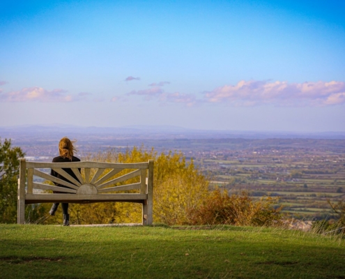 A person sitting alone on a wooden bench on a grassy hill, looking out over a wide valley under a clear evening sky