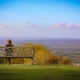 A person sitting alone on a wooden bench on a grassy hill, looking out over a wide valley under a clear evening sky