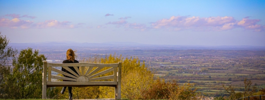 A person sitting alone on a wooden bench on a grassy hill, looking out over a wide valley under a clear evening sky
