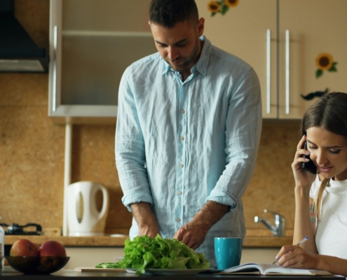Woman on a phone call taking notes at a kitchen counter while her partner prepares a salad
