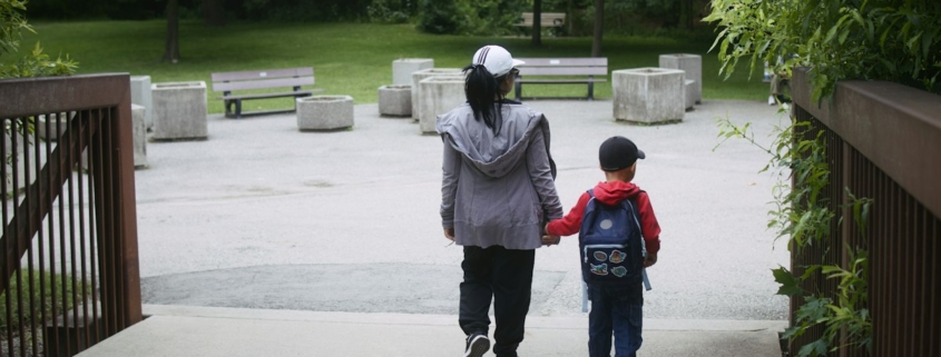A parent and young child walk hand in hand along a tree-lined park path