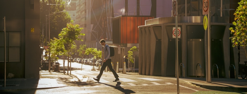 A person with a backpack walking across a quiet city street in early morning light on the way to work