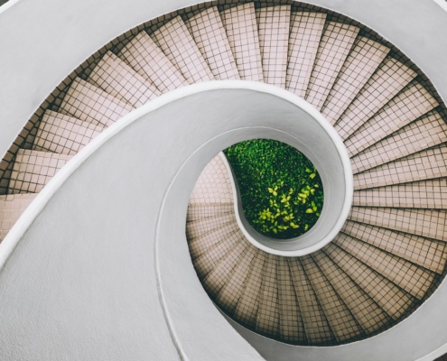 A bright white spiral staircase seen from above, curving inward toward green plants growing at its center.