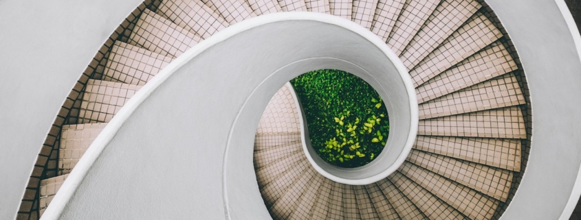 A bright white spiral staircase seen from above, curving inward toward green plants growing at its center.