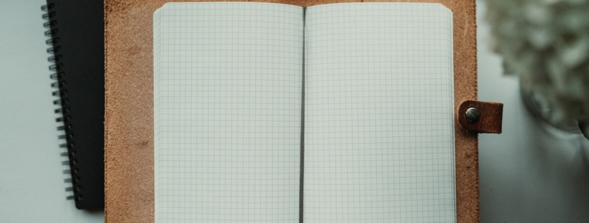 Open blank grid-paper journal in a leather cover on a desk beside white flowers, for keeping a daily medication and mood log