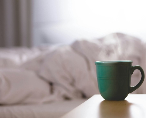 A green ceramic mug on a bedside table beside an unmade bed in soft morning light