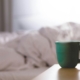 A green ceramic mug on a bedside table beside an unmade bed in soft morning light